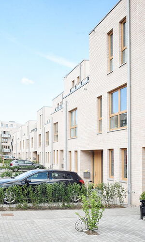 Moderne Reihenhäuser aus hellem Backstein mit großen Fenstern und Holztüren an einer breiten, gepflasterten Straße unter blauem Himmel