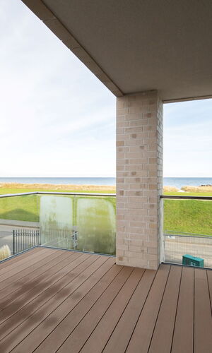 Balkon mit Holzterrasse und Glasgeländer, Blick auf eine grüne Wiese und das Meer am Horizont
