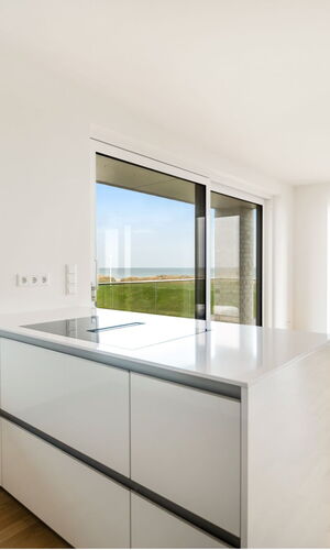 Helle, moderne Küche mit weißer Kücheninsel und Blick durch große Fenster auf eine grüne Wiese und den Horizont